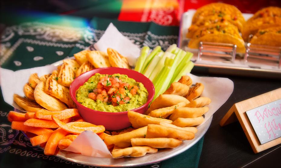 Avocado hummus, pita and carrots displayed played on a plate with tacos in the background. 